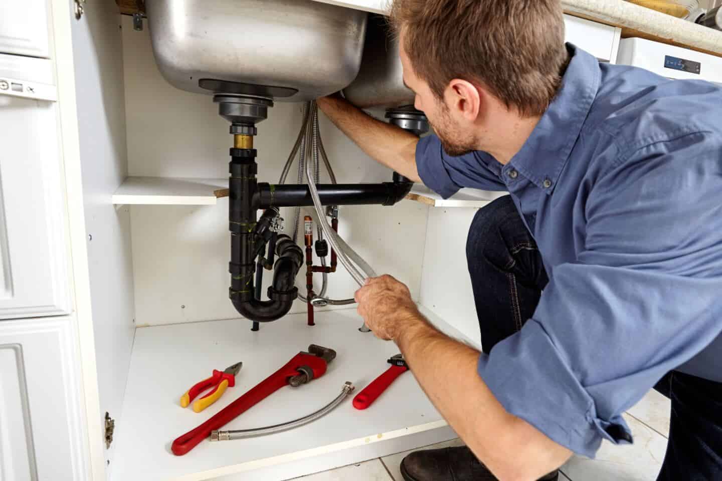 A plumber kneels below a sink and checks the pipework