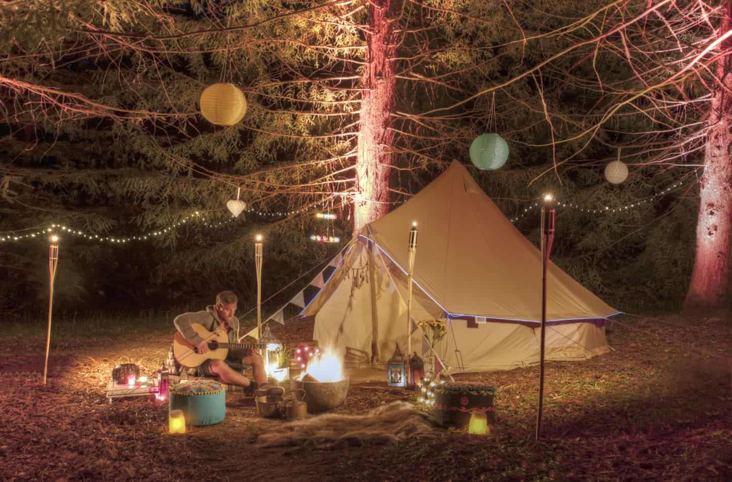 A man sat outside a tent in a woodland clearing playing guitar surrounded by tiki torches and paper lanterns hanging from trees.