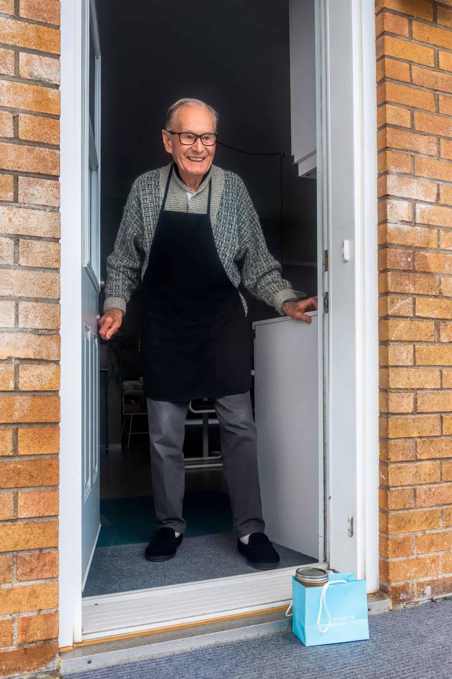 An smiling old man stands inside his open front door. There is a package on the step that has been left for him