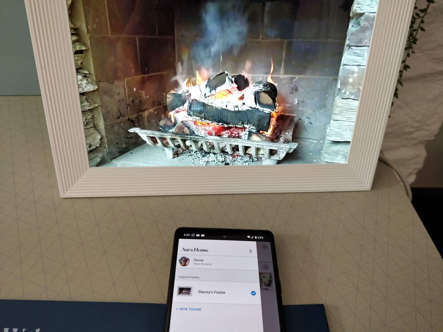 A digital photo frame on a desk displaying a video of a roaring fire. A mobile phone on the desk displays the app that is linked to the digital photo frame