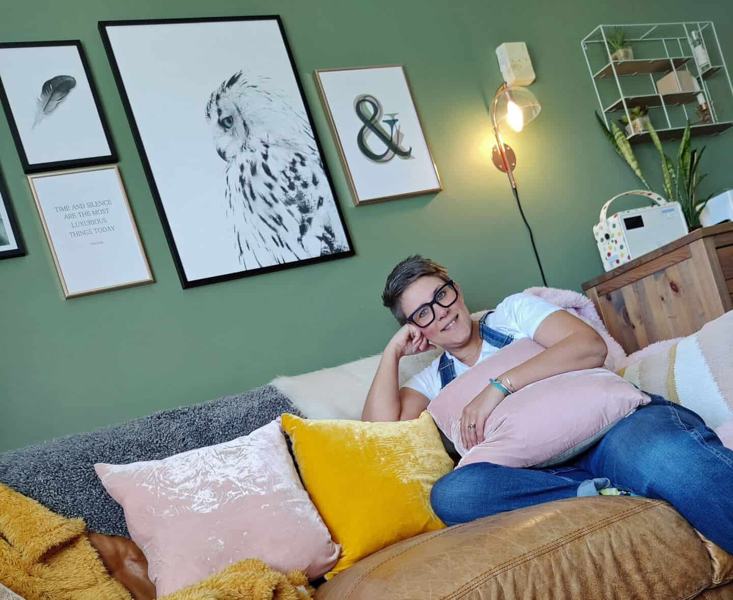 A middle aged woman sits in a green living room on a brown leather sofa with artwork above her head. There are pink and yellow cushions on the sofa.