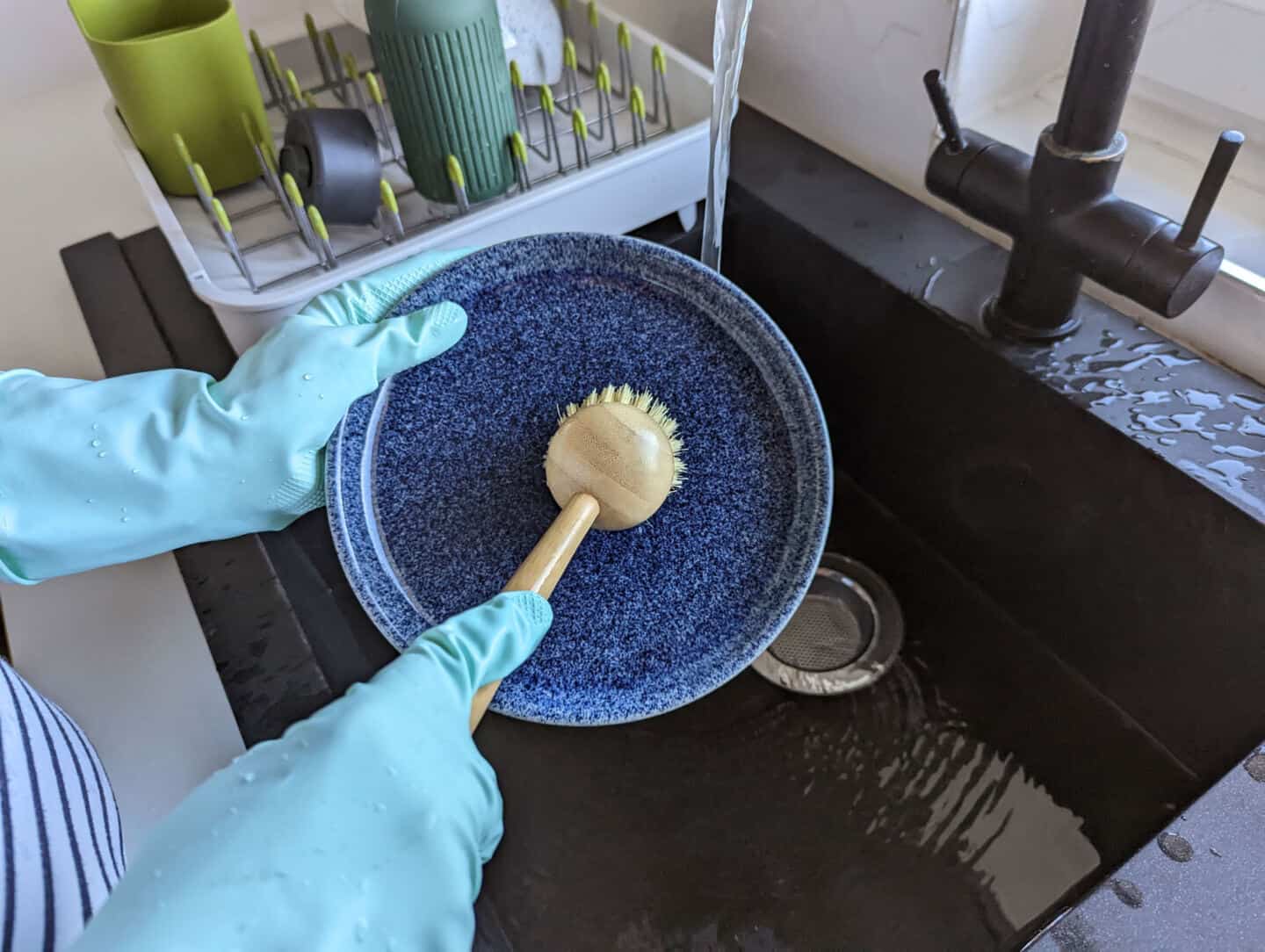 A bamboo washing up brush by Seep is used to clean a blue Denby plate over a black granite sink