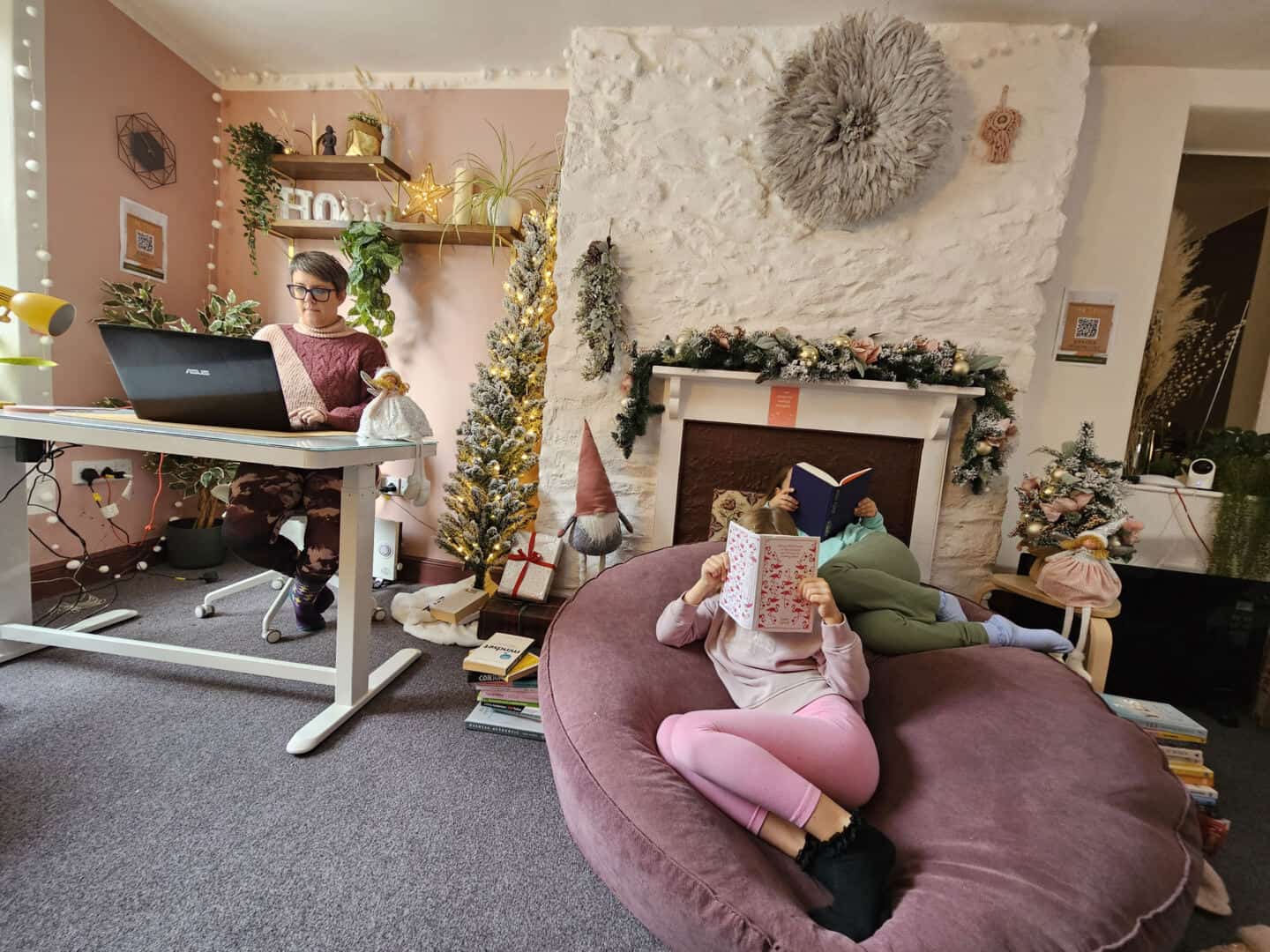 Two small girls sat reading on a bean bag while a woman in her forties works on a laptop at a desk nearby