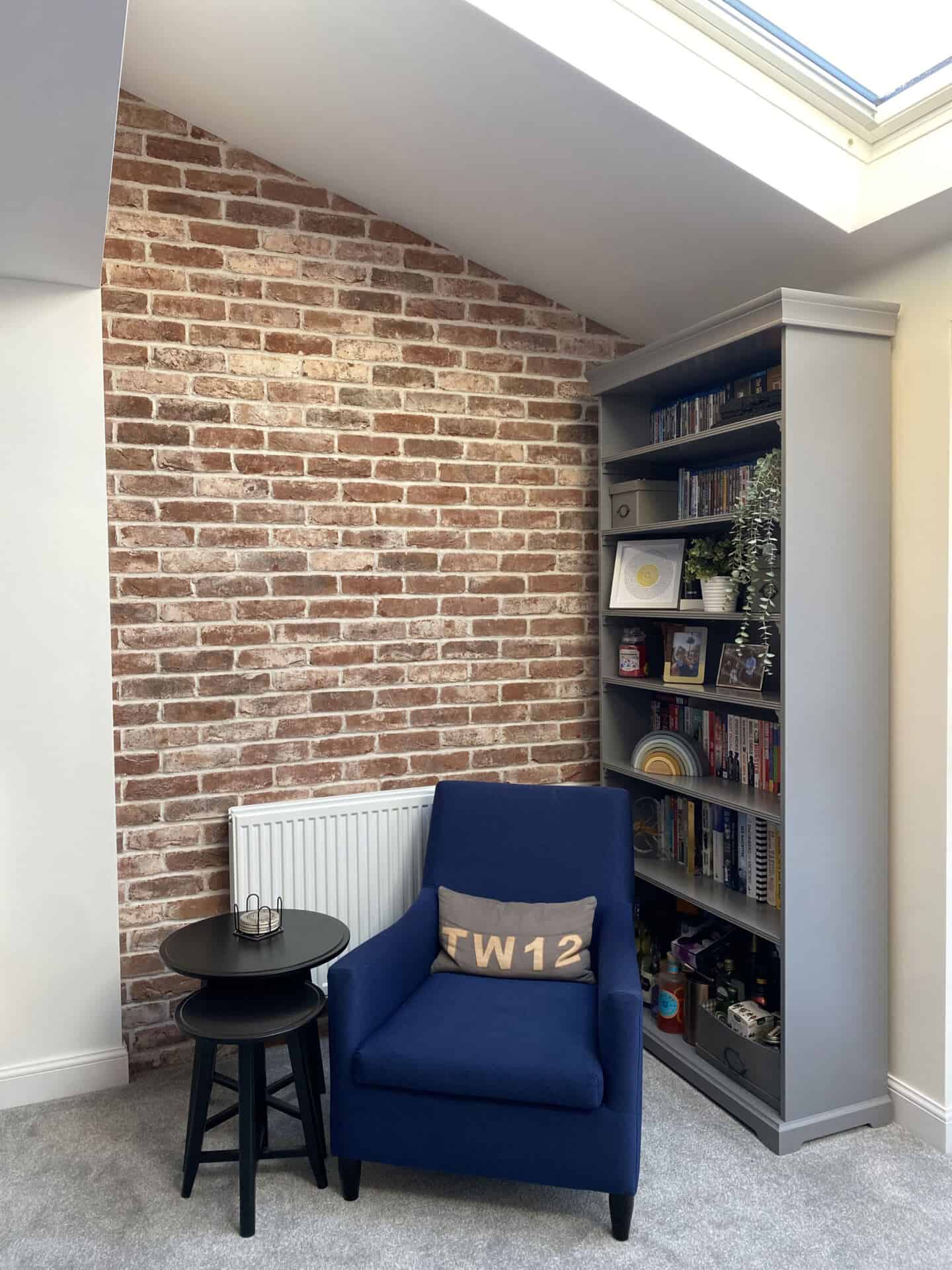 A reading nook with cosy chair and small black side tables under the roof eaves. A tall bookcase full of books is against the wall behind the chair.