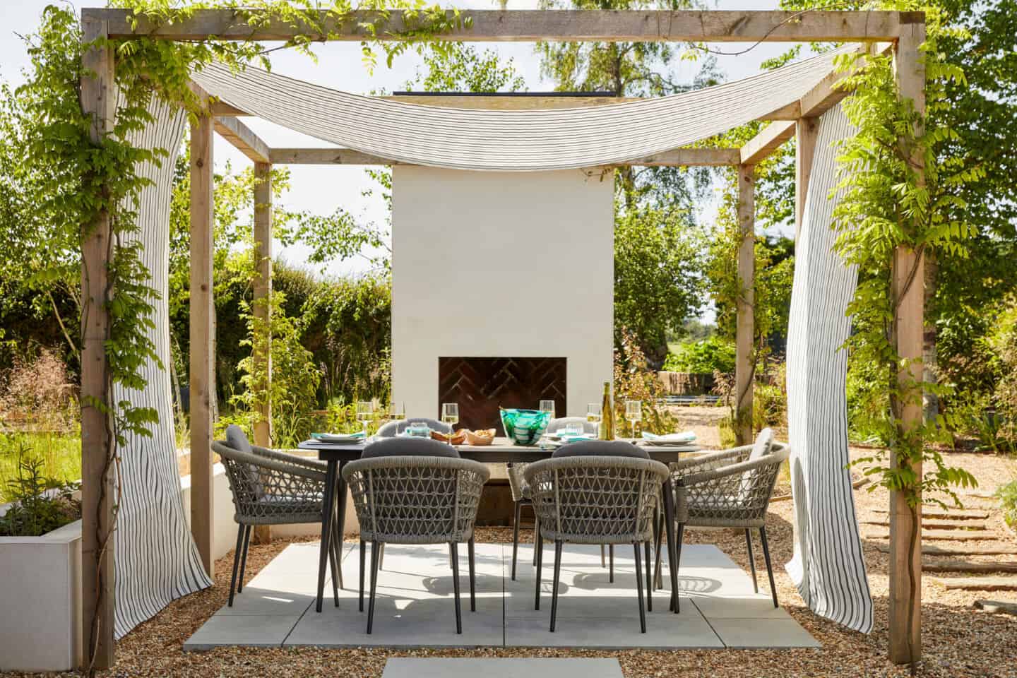 A large garden featuring an outdoor dining scene beneath a pergola draped with white fabric