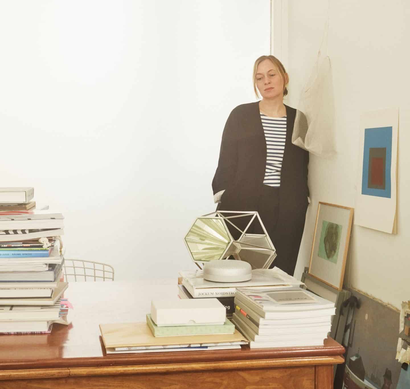 Danish designer Cecilie Manz standing behind a desk piled high with books.