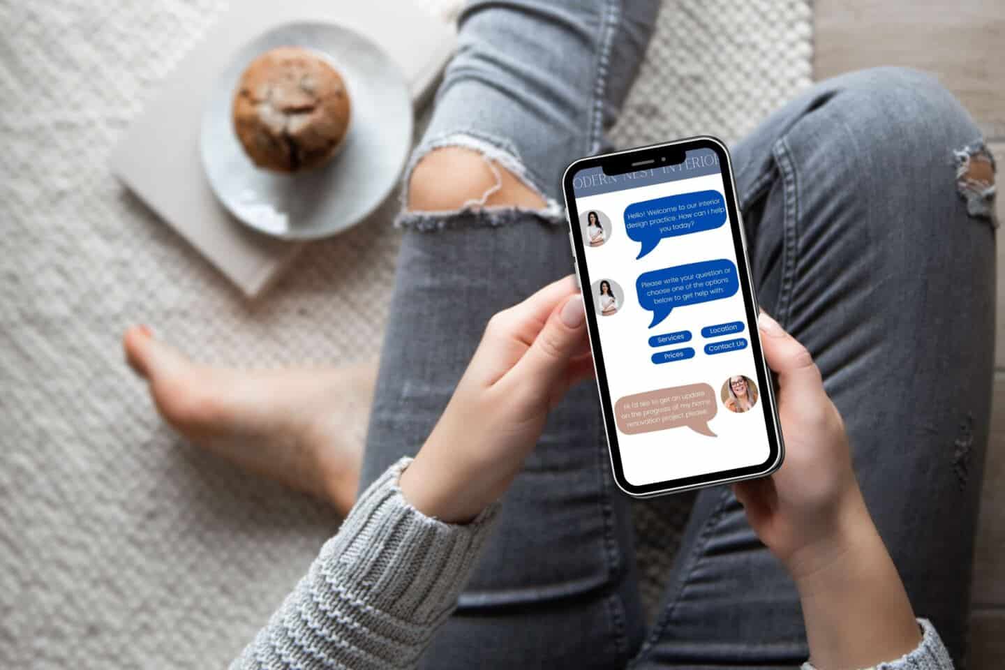 A woman sat crossed legged on the carpet holding a smart phone. The screen shows her talking to a chatbot at an interior design company