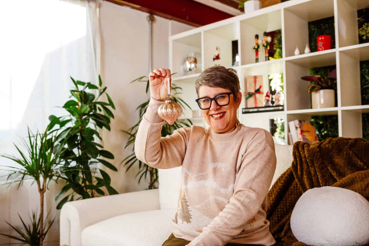 A woman in her forties sat on a sofa  holding up a Christmas bauble from Balsam Hill 