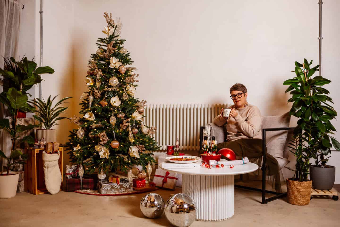 A woman in her forties sat on a chair beside a realistic artificial Christmas tree drinking hot chocolate