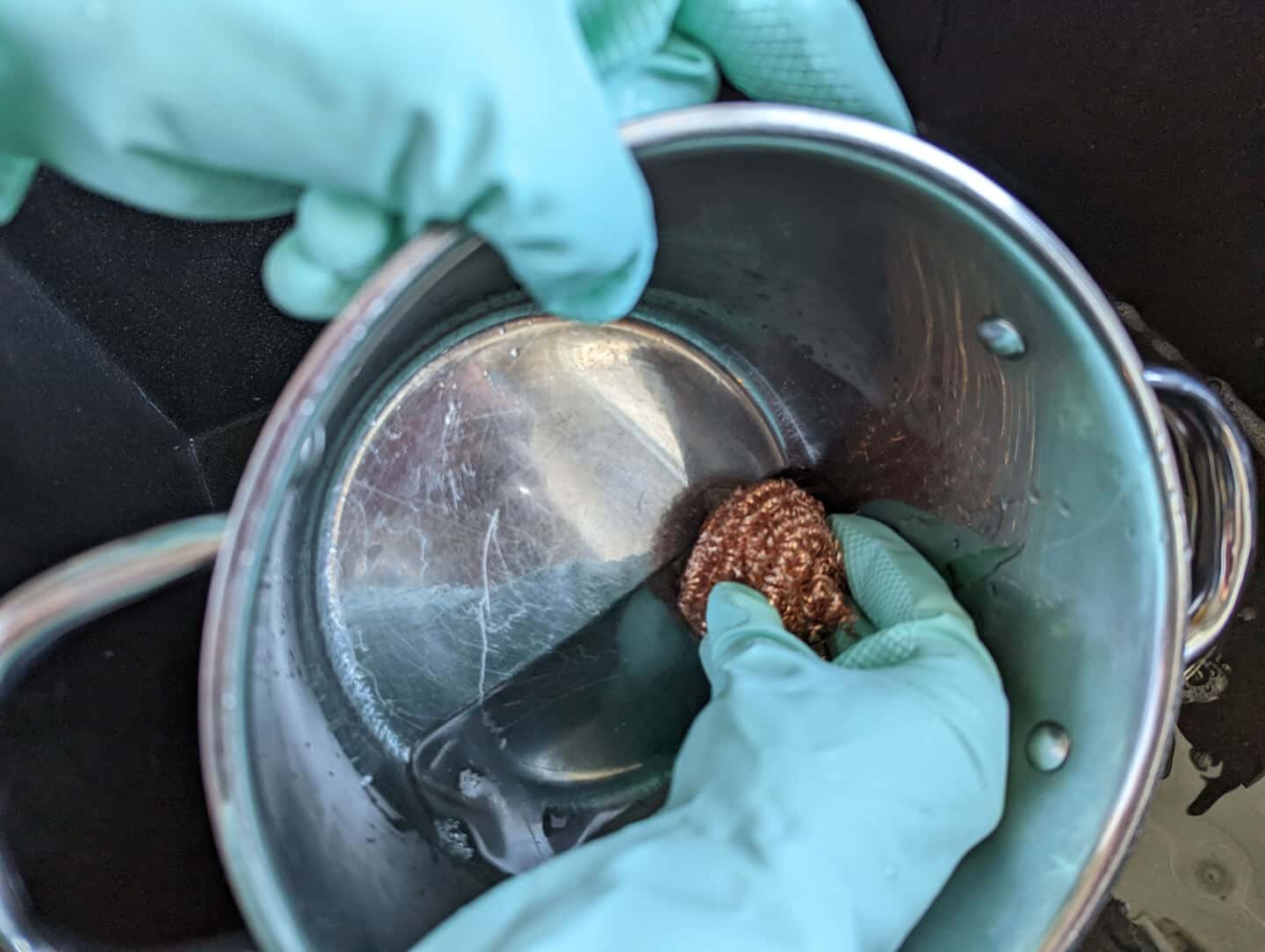 A copper scourer by Seep being used to clean a large steel saucepan. The hand wears natural rubber gloves by Seep