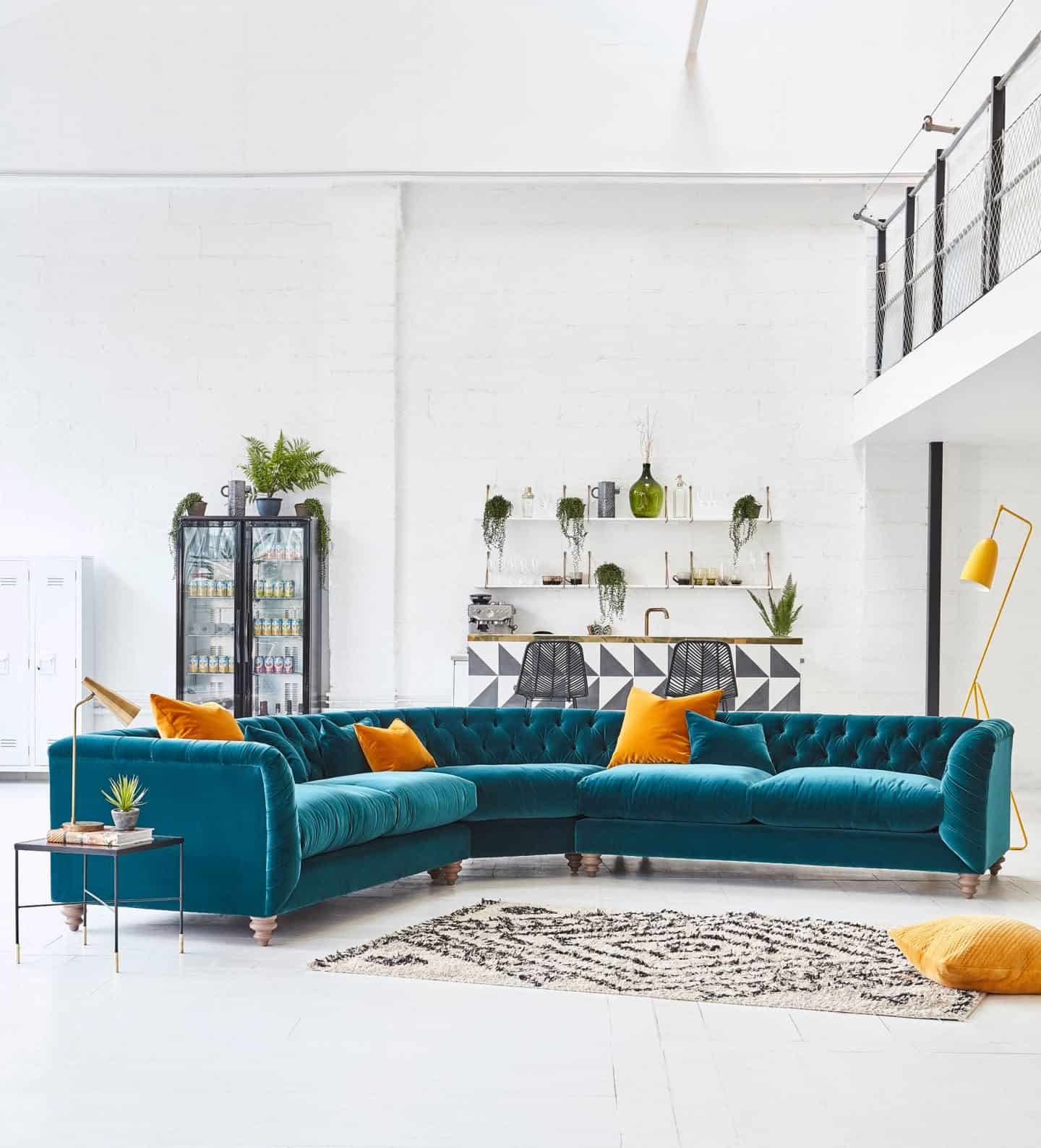 A large turquoise velvet corner sofa  from Darlings of Chelsea with mustard yellow scatter 
cushions sits in a warehouse setting. Furniture can be found on furniture comparison website ufurnish.com