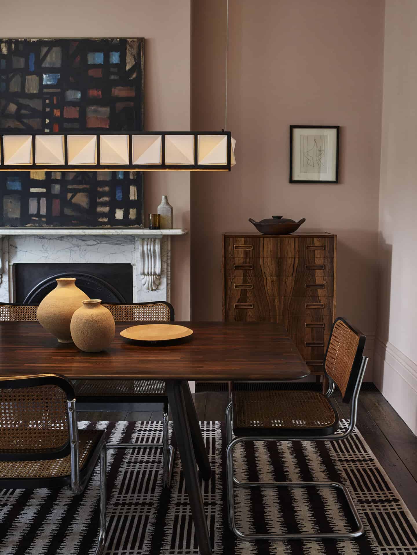 A dining room with mushroom coloured walls, a dark wood dining table and rattan chairs. A chest of drawers is in the corner of the room next to the marble fireplace. A large square long light hangs above the table.