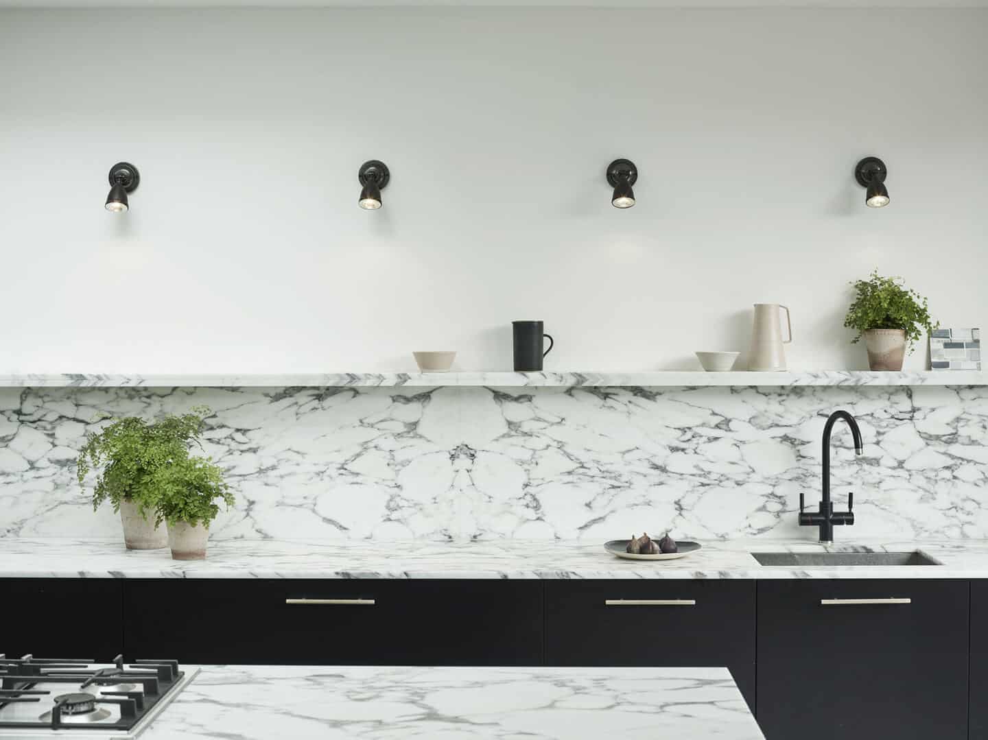 A navy blue kitchen wit marble worktop and back splash. Four spot lights on the wall above