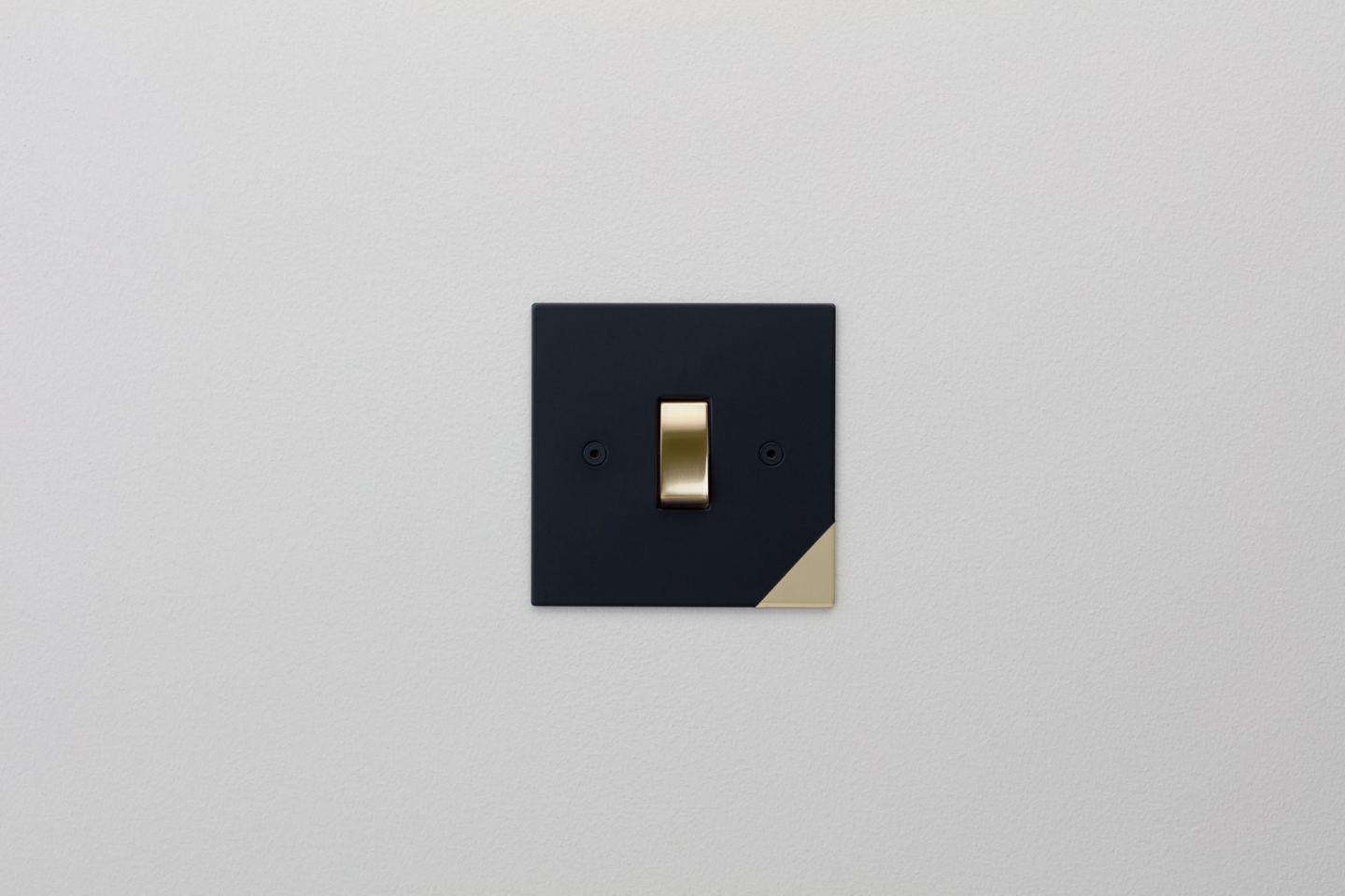 Designer light switches by Kelly Hoppen for Focus SB. Corner is a matt black switch plate with a gold switch and a subtle gold triangle in the bottom right-hand corner. 