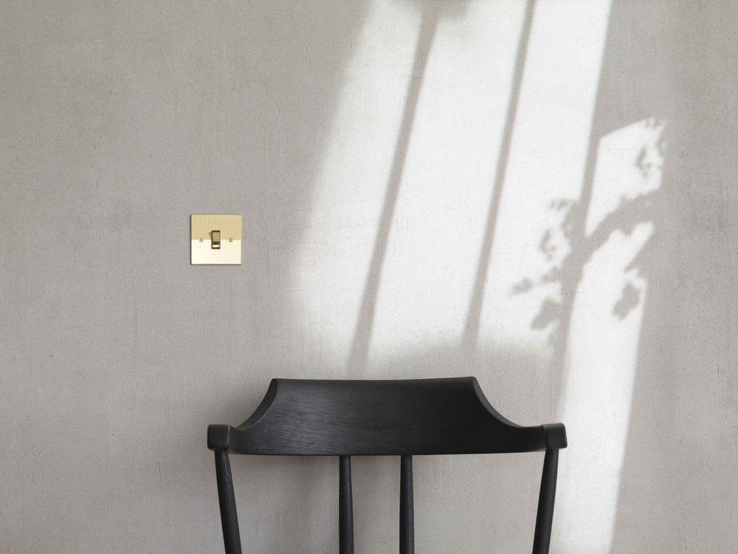 Designer switches and sockets by Kelly Hoppen for Focus SB. A gold light switch on a grey wall above a black wooden chair.