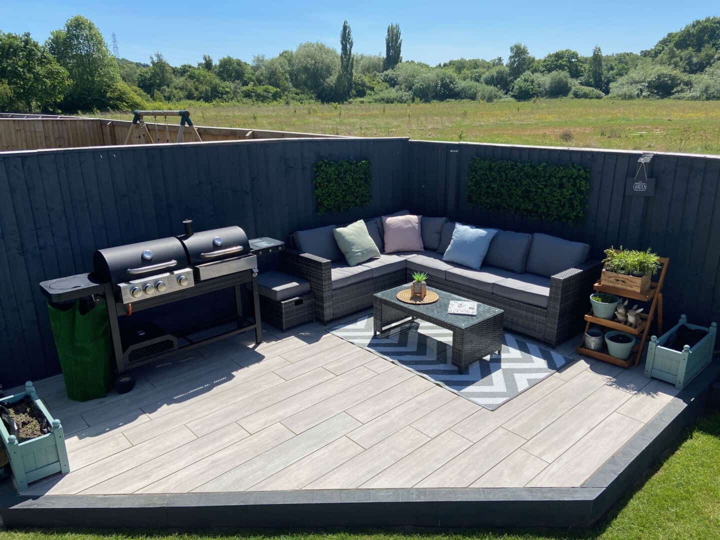 A low-maintenance garden with tiles deck in the corner with sofa, BBQ and coffee table surrounded by grey fences