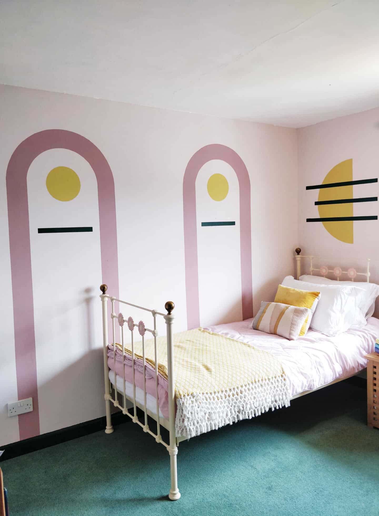 A single bed with pastel pink bedding and geometric pink and yellow cushions. There are painted arches and geometric shapes on the walls in pink, green and yellow.