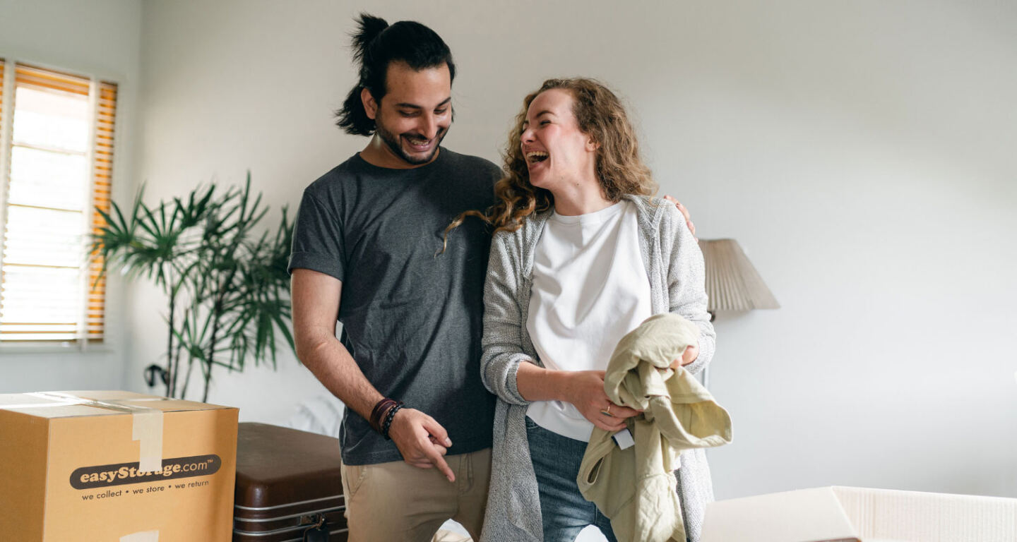 A young couple laugh while packing belongings into cardboard boxes 