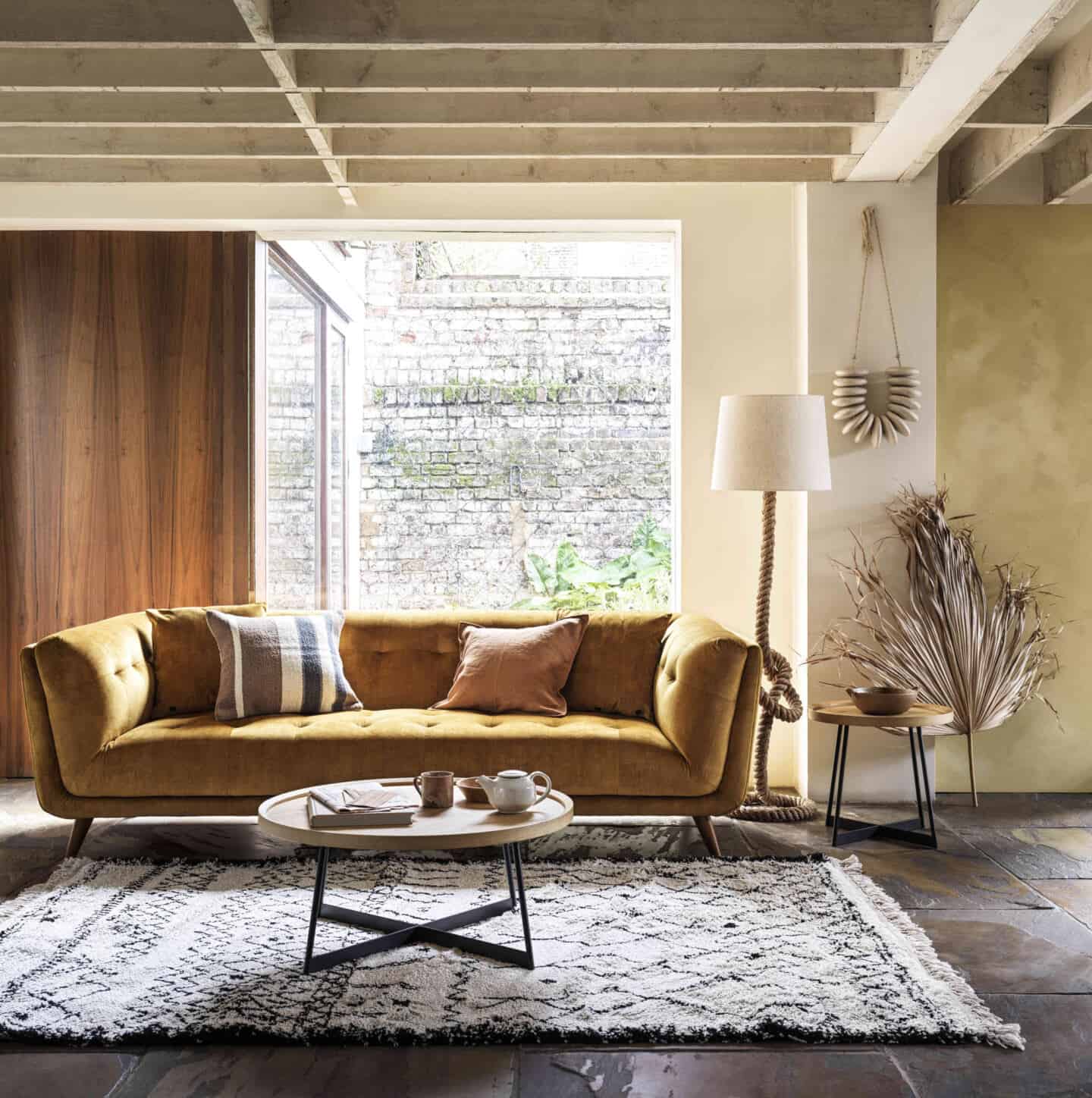 An ochre tuxedo sofa with berber rug and round coffee table in front and large window behind.