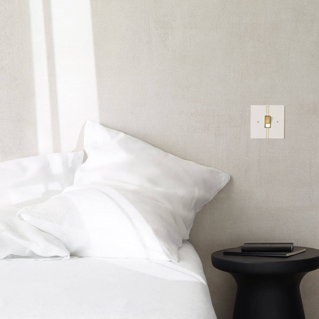 Designer light switches by Kelly Hoppen for Focus SB. A white light switch on a grey wall beside a bed made up with crisp white bed linen. 