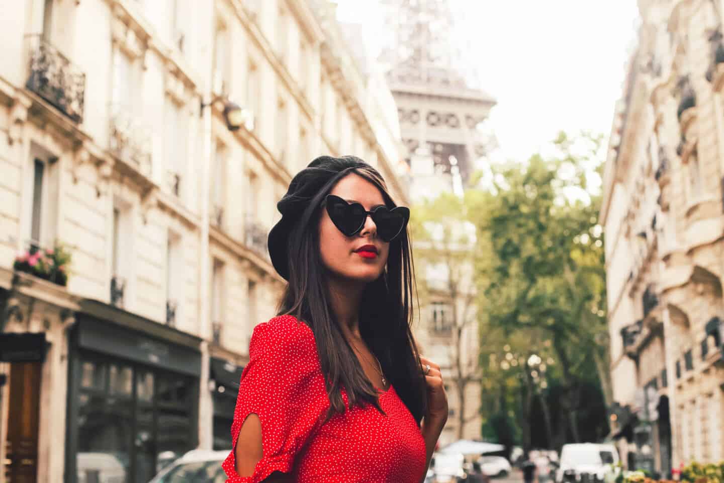 A young woman in red dress, black beret and heart-shaped sunglasses stood in a Parisian street with a view of the Eiffel Tower behind