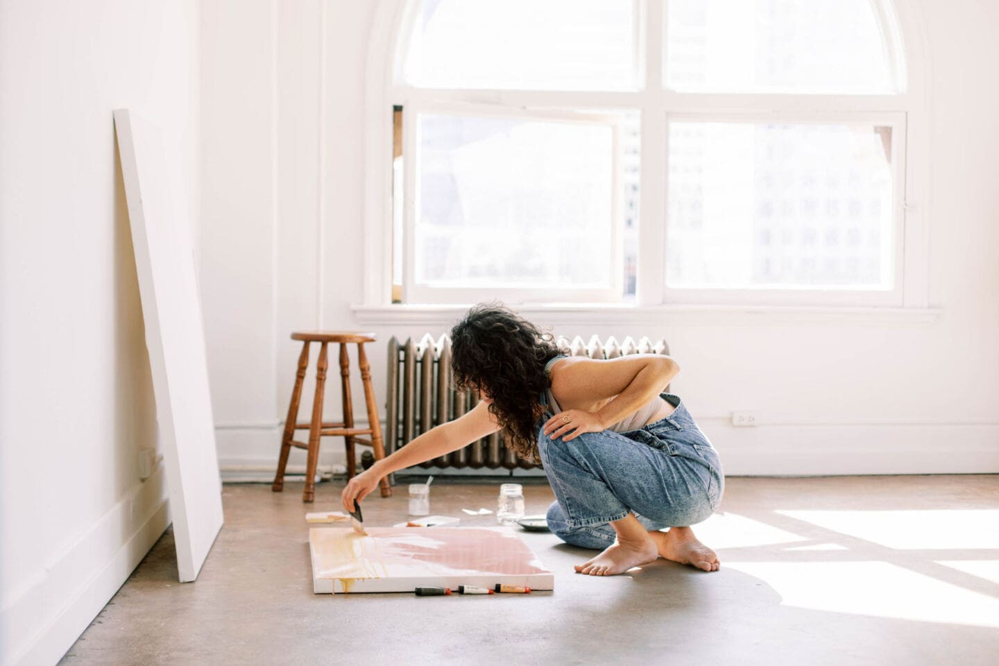 A young woman paints onto large canvas on the floor of a white apartment room