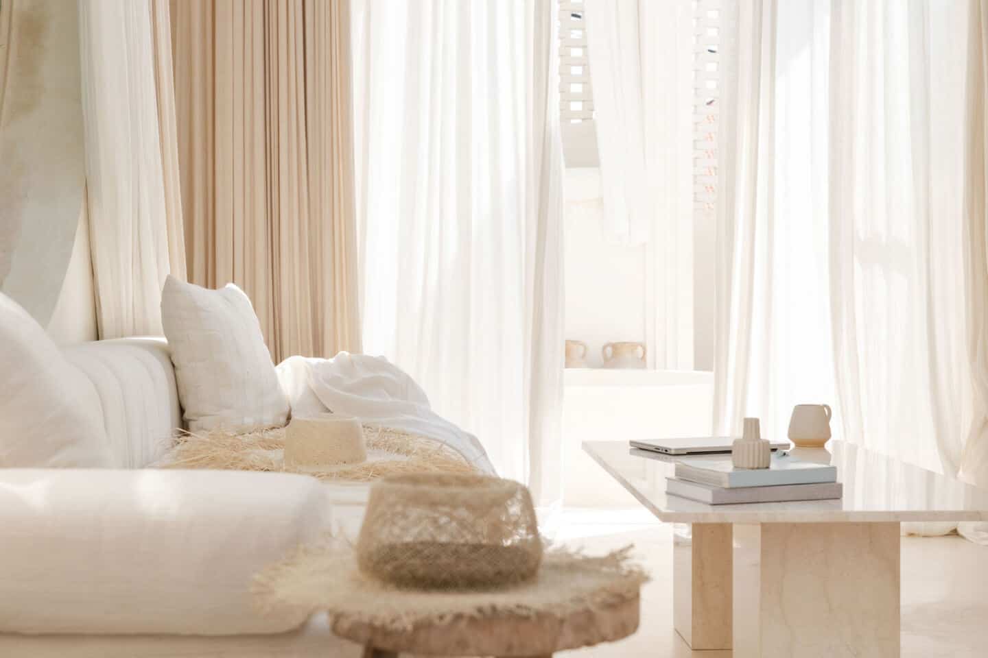 A neutral living room with floaty fabrics on the windows billowing in the wind, a white linen sofa and a beige marble coffee table 