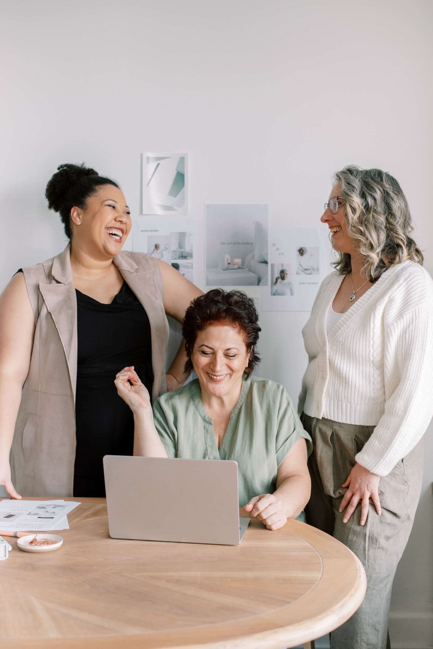 Two middle aged women standing and laughing behind another middle aged woman sat at a desk looking at a laptop