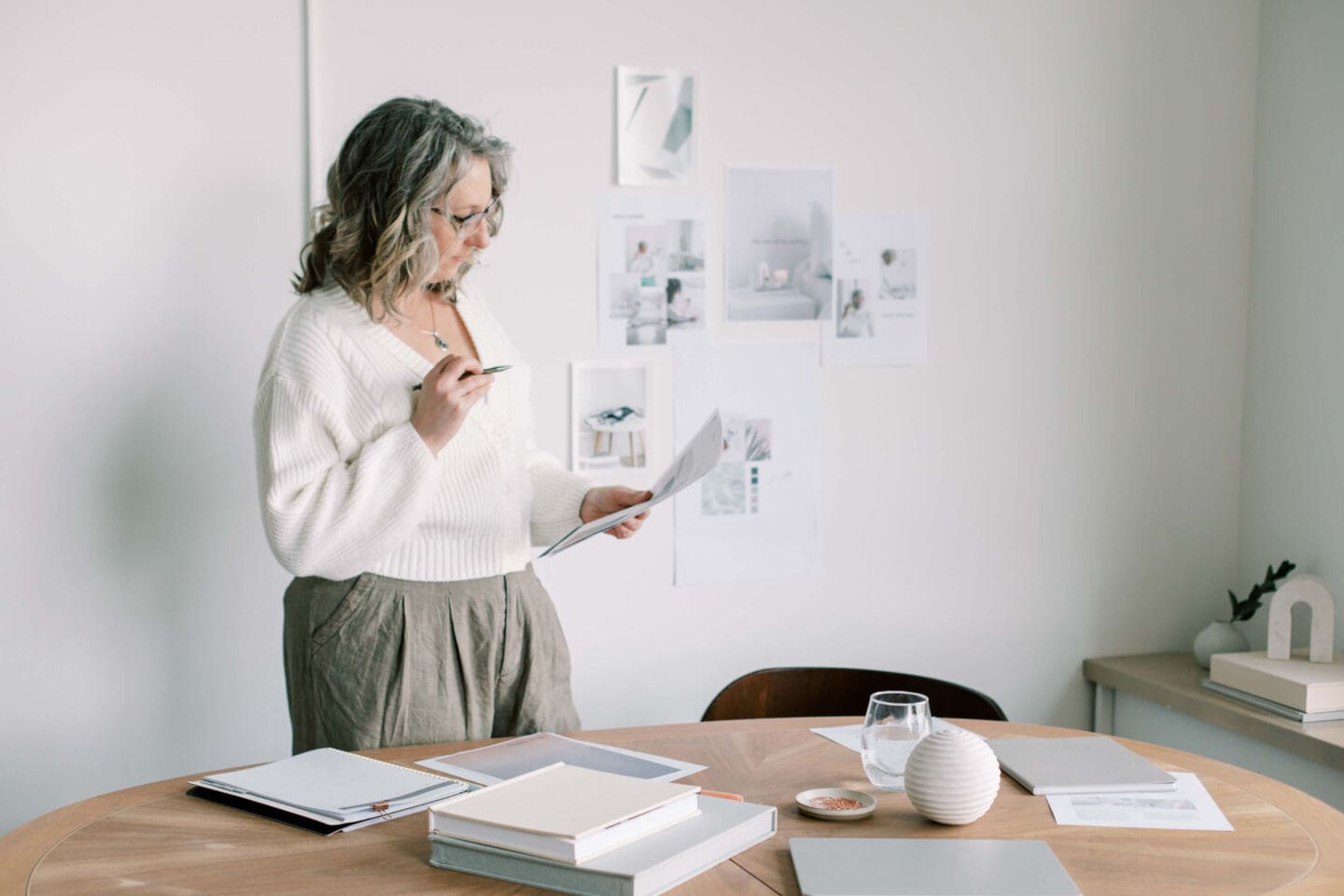 A woman stands by a desk looking at a piece of paper and holding a pen