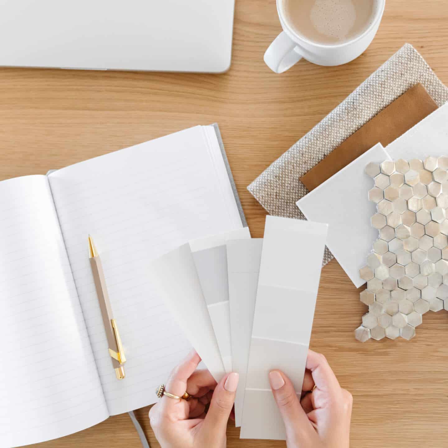 A woman's hands hold paint chits above a notebook and next to fabric and tile samples for an interior design project