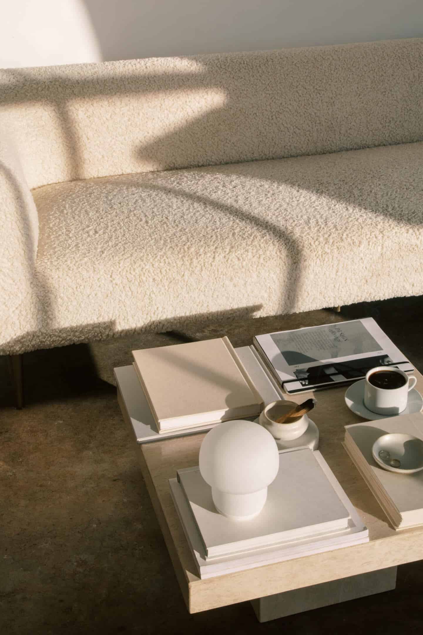 A minimal monochrome living room featuring a low-level coffee table next to a boucle sofa. There are books, a lamp and a coffee on the table.