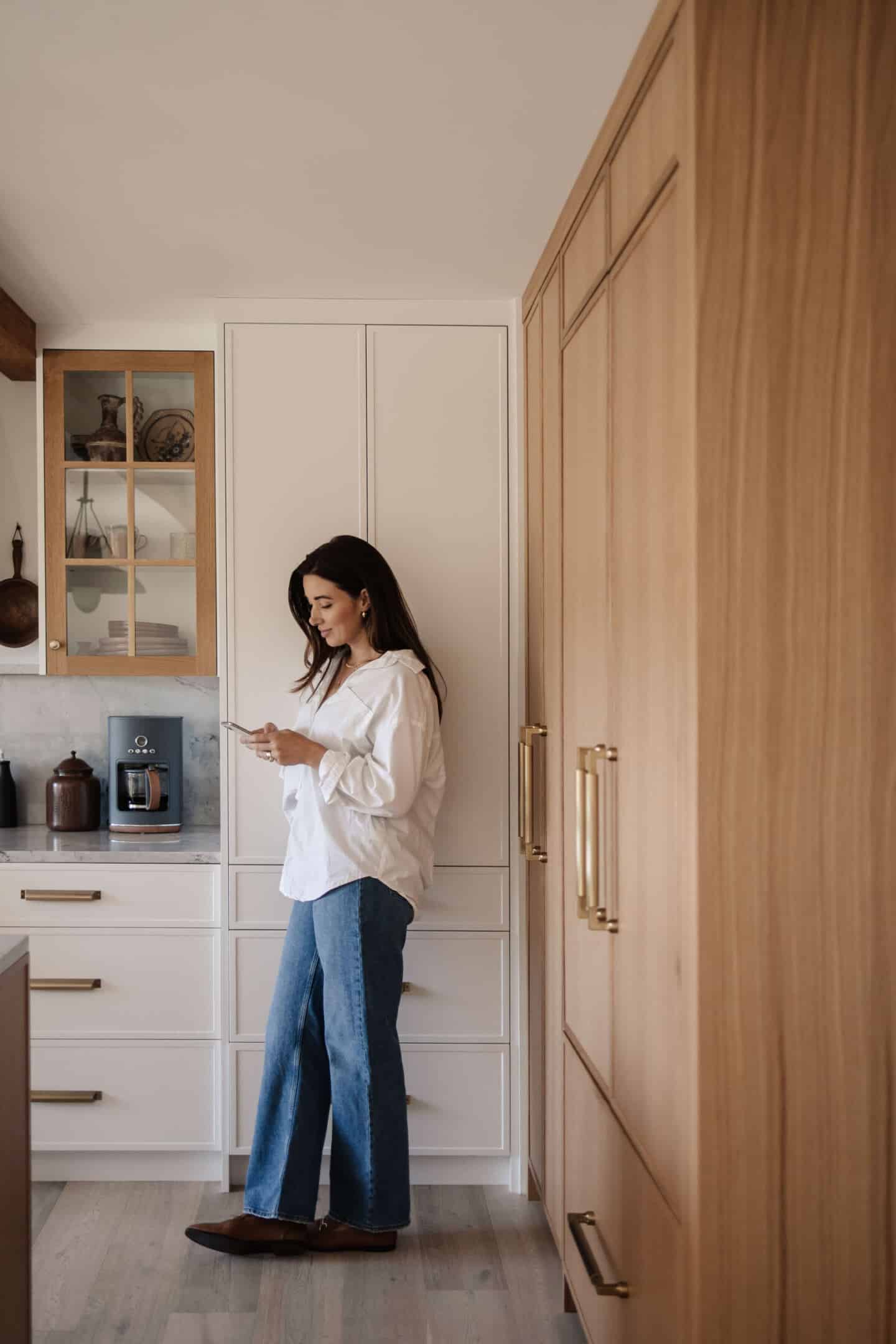 A young woman wearing a white shirt and jeans stands in a kitchen looking at her mobile phone