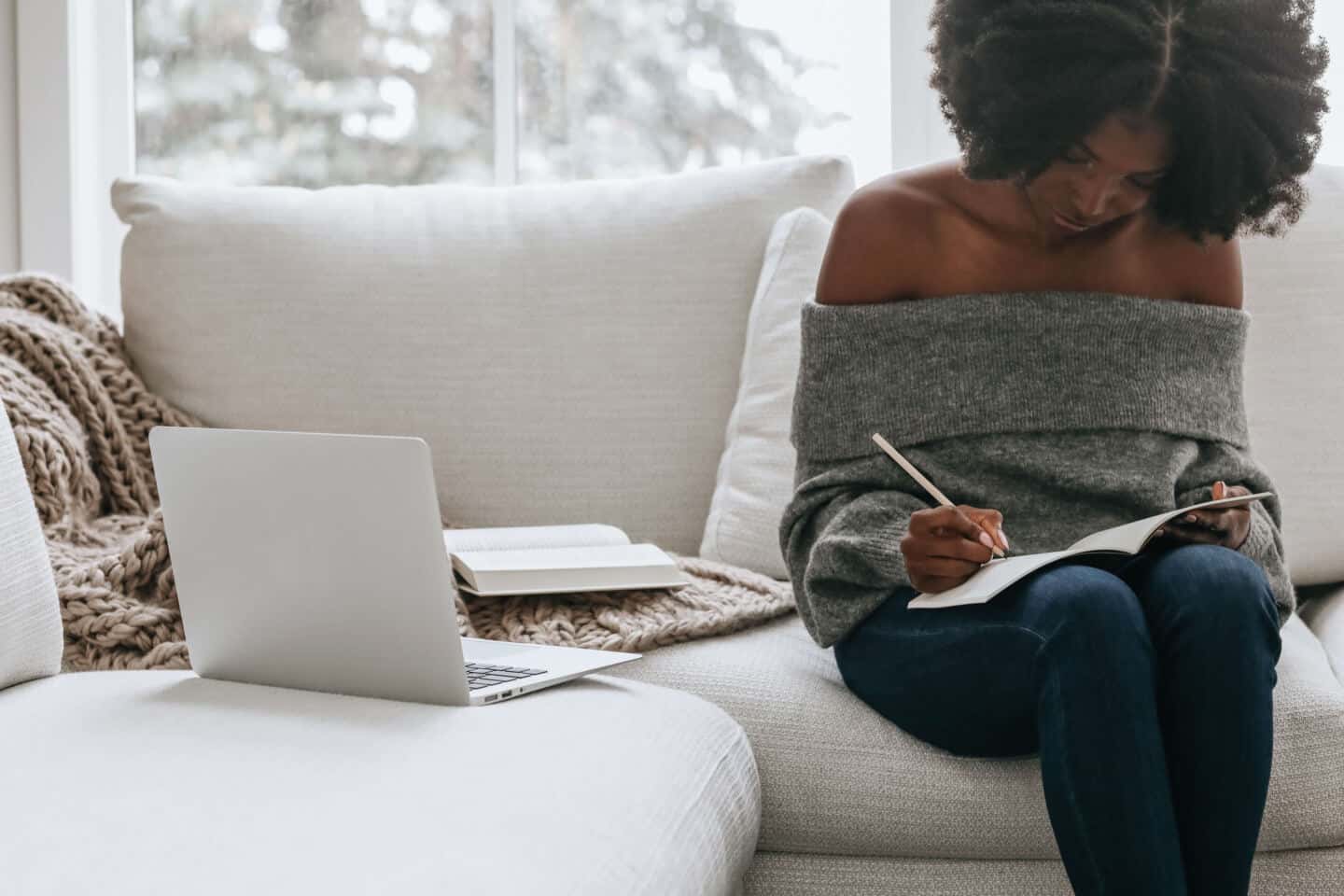 A young black woman sat on a grey sofa writing in a notebook. A laptop and an open book are on the sofa next to her.