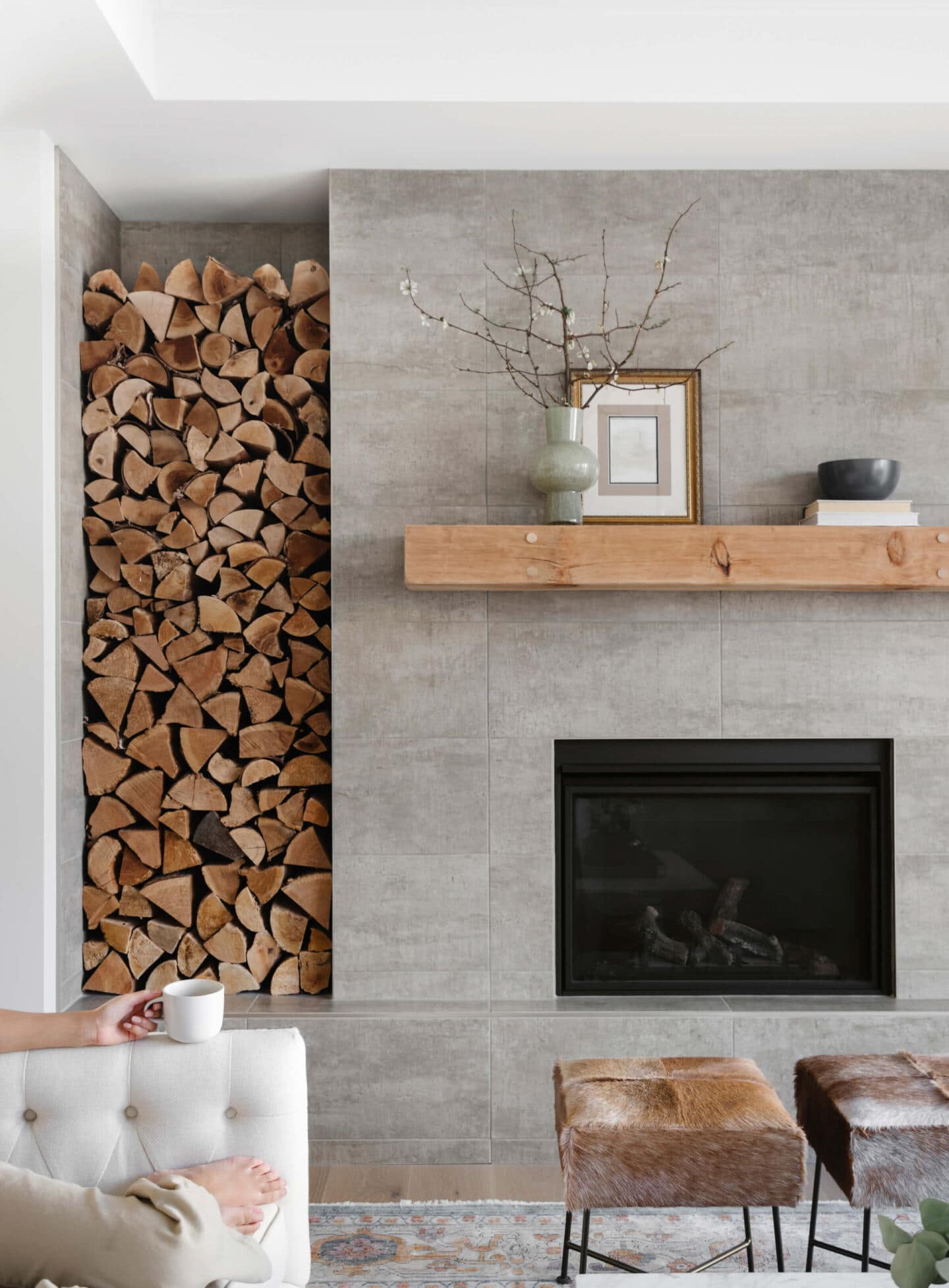 A fireplace in a luxury home with a large wooden beam above and chopped wood piled in an alcove to the left of the fire