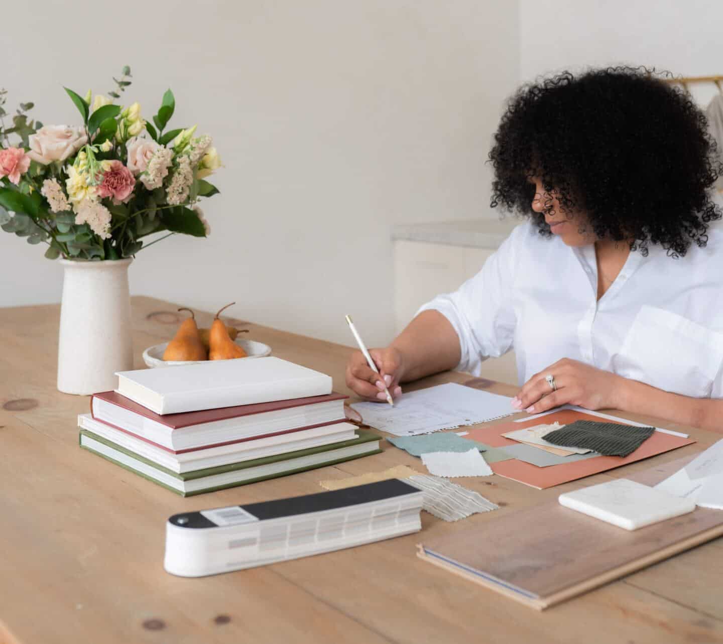 A woman sat at a desk designing a home. She is surrounded by fabric samples and colour swatches and is drawing the floor plan of a room