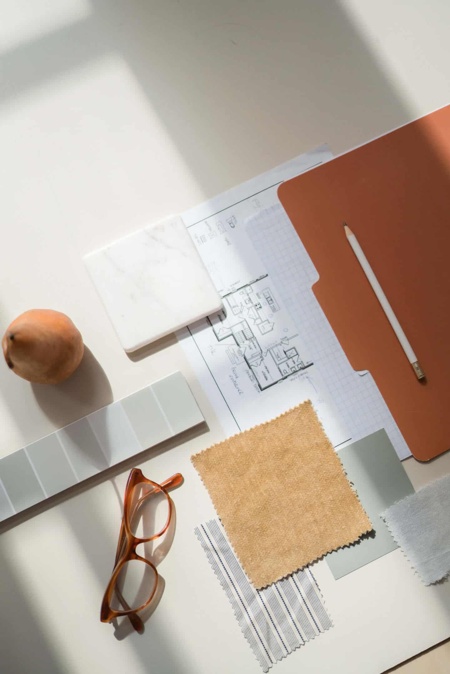 An architectural plan on desk next to fabric samples, a paint chit, a tile sample and a pair of glasses