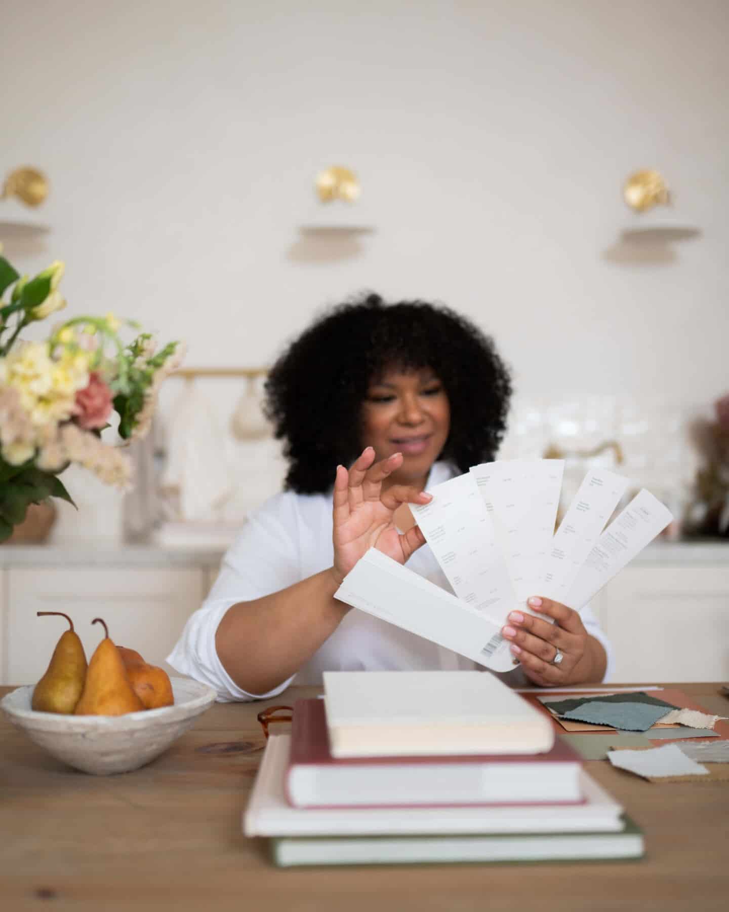 A young black woman who runs an interior design business sits at a dining room table covered in fabric swatches and looks through a fan of paint colours. 