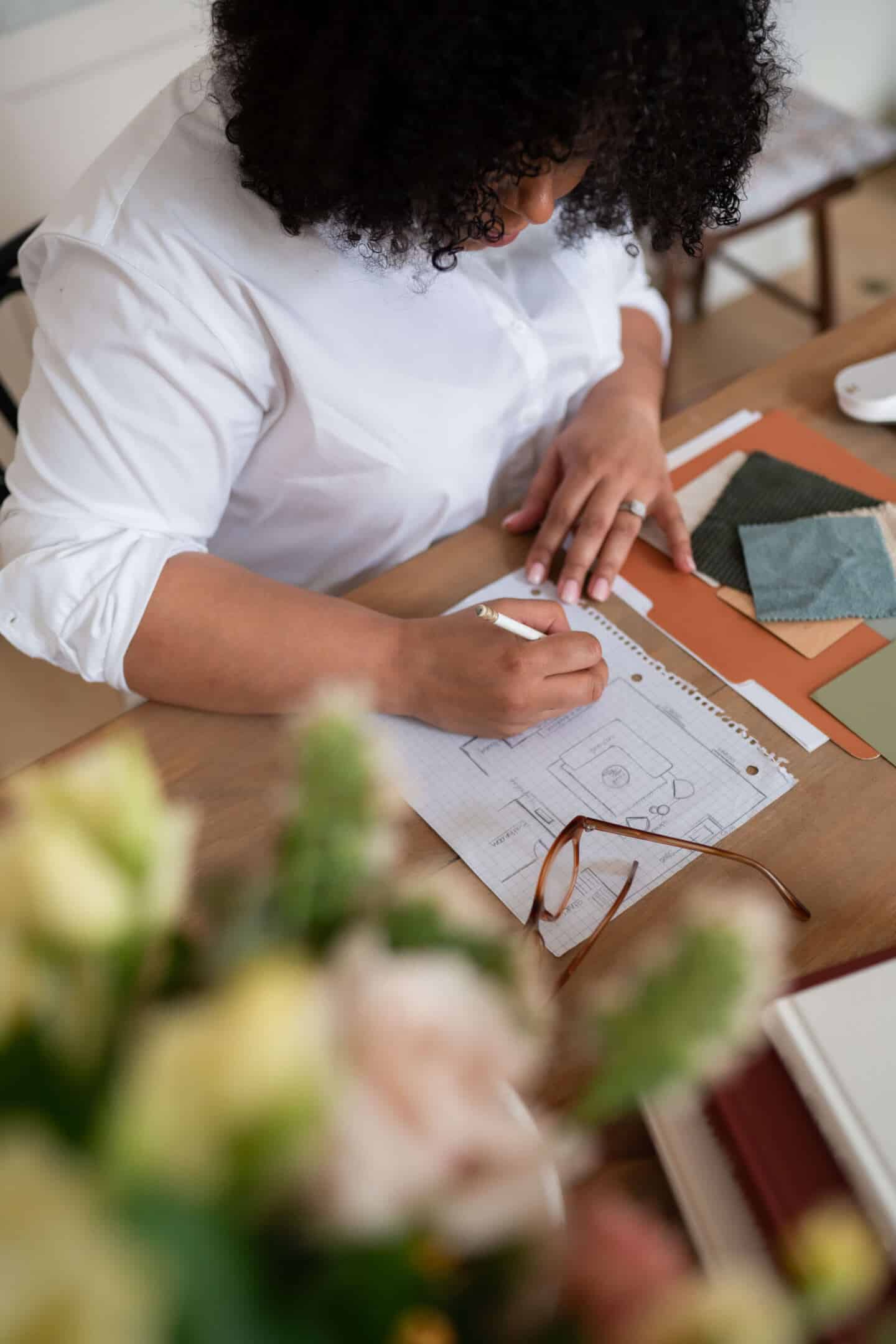 A woman sat at a desk designing a home. She is surrounded by fabric samples and colour swatches and is drawing the floor plan of a living room