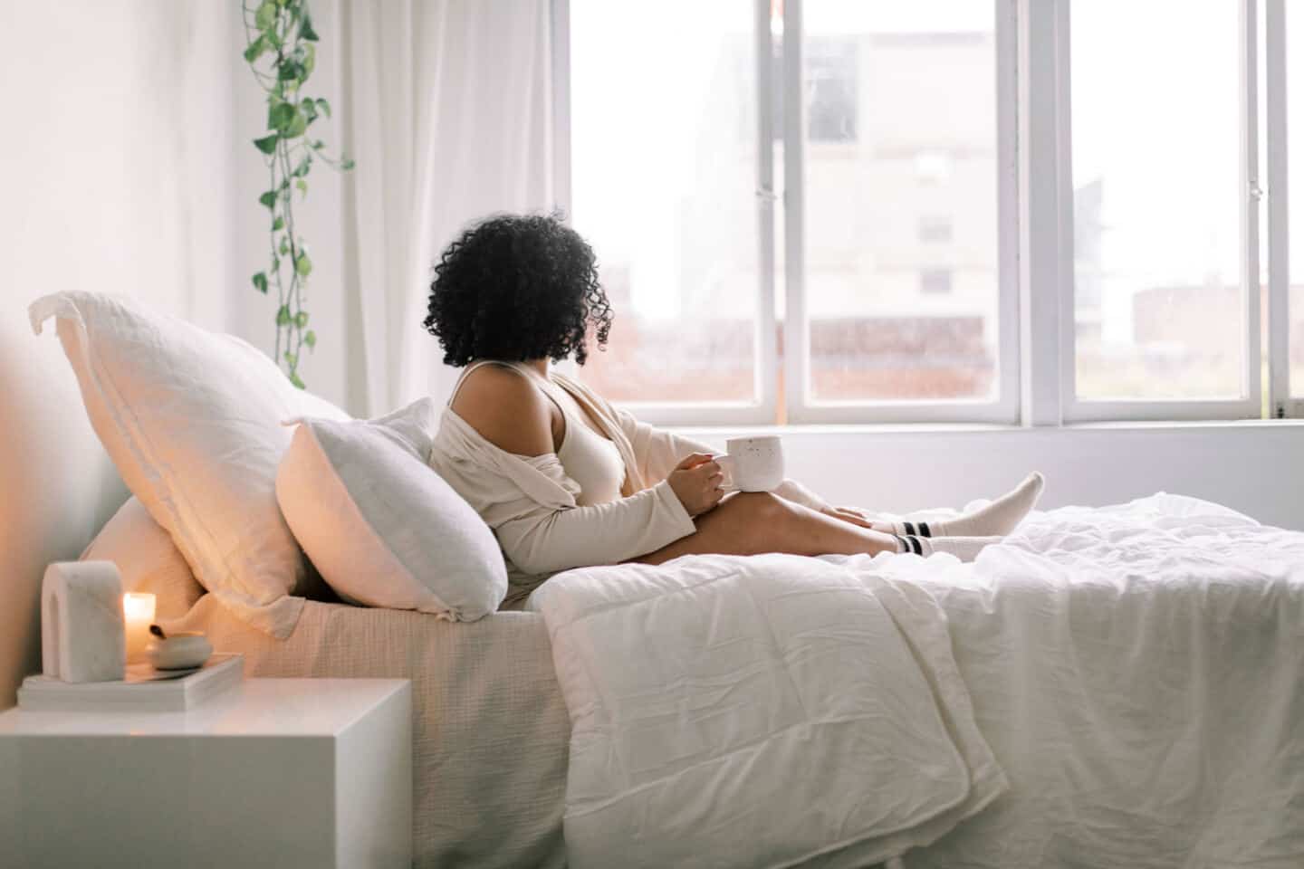 A young woman sat in a bed in oyjamas and socks holding a mug and looking out the window