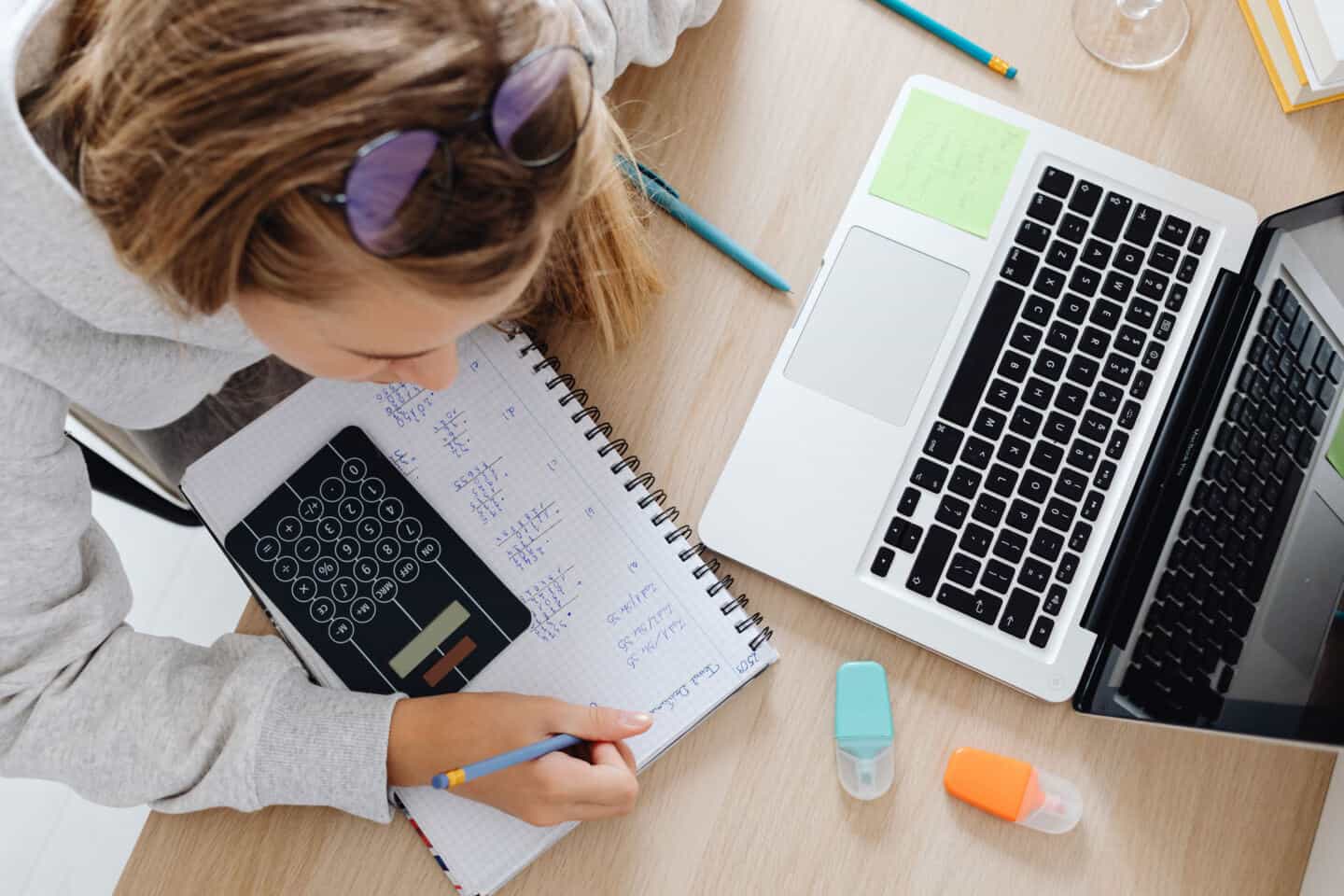 A woman sat at a laptop writing in a notebook and using a calculator