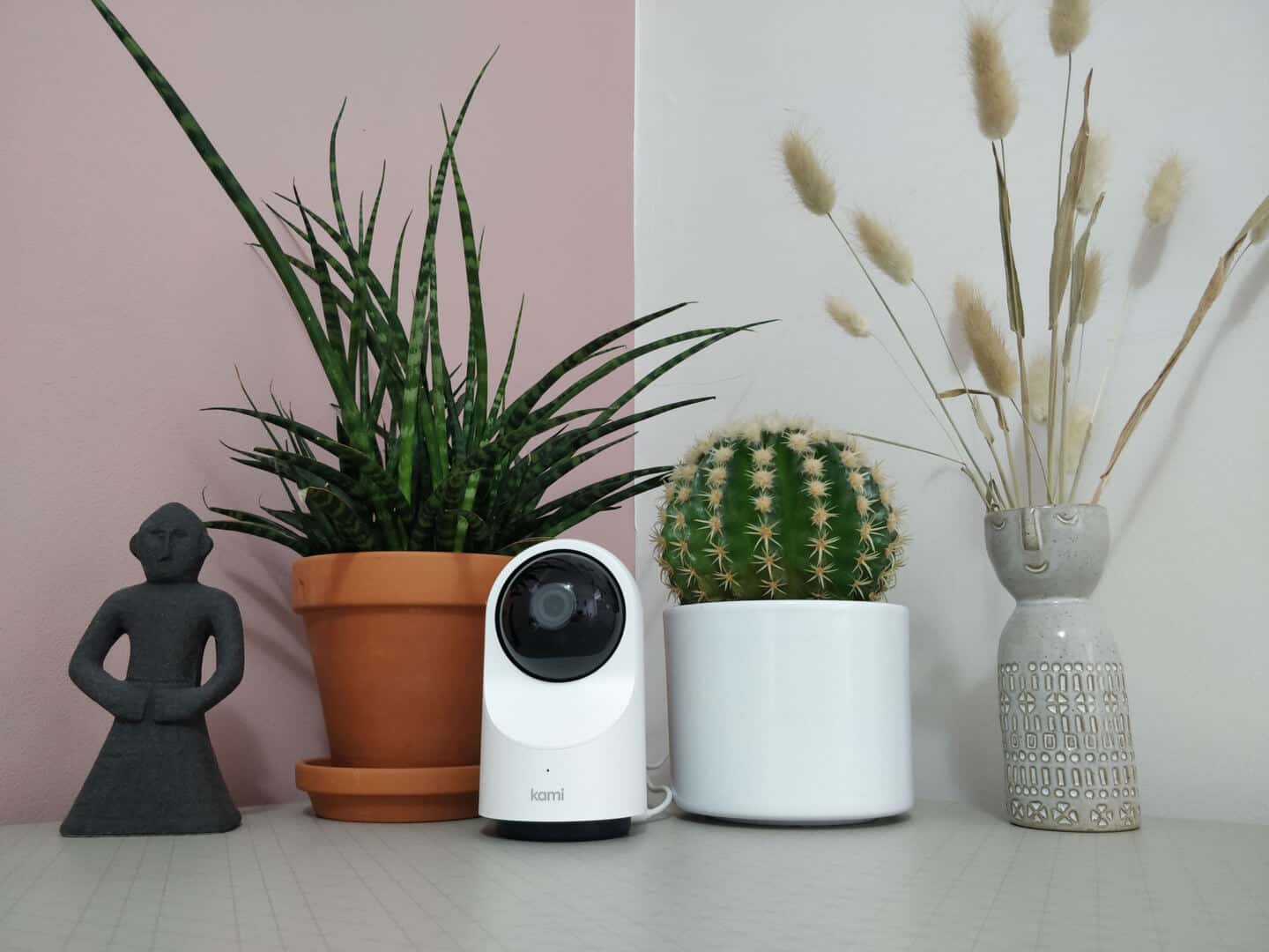 Kami indoor home security camera on a table between two plants and vase and a statue.