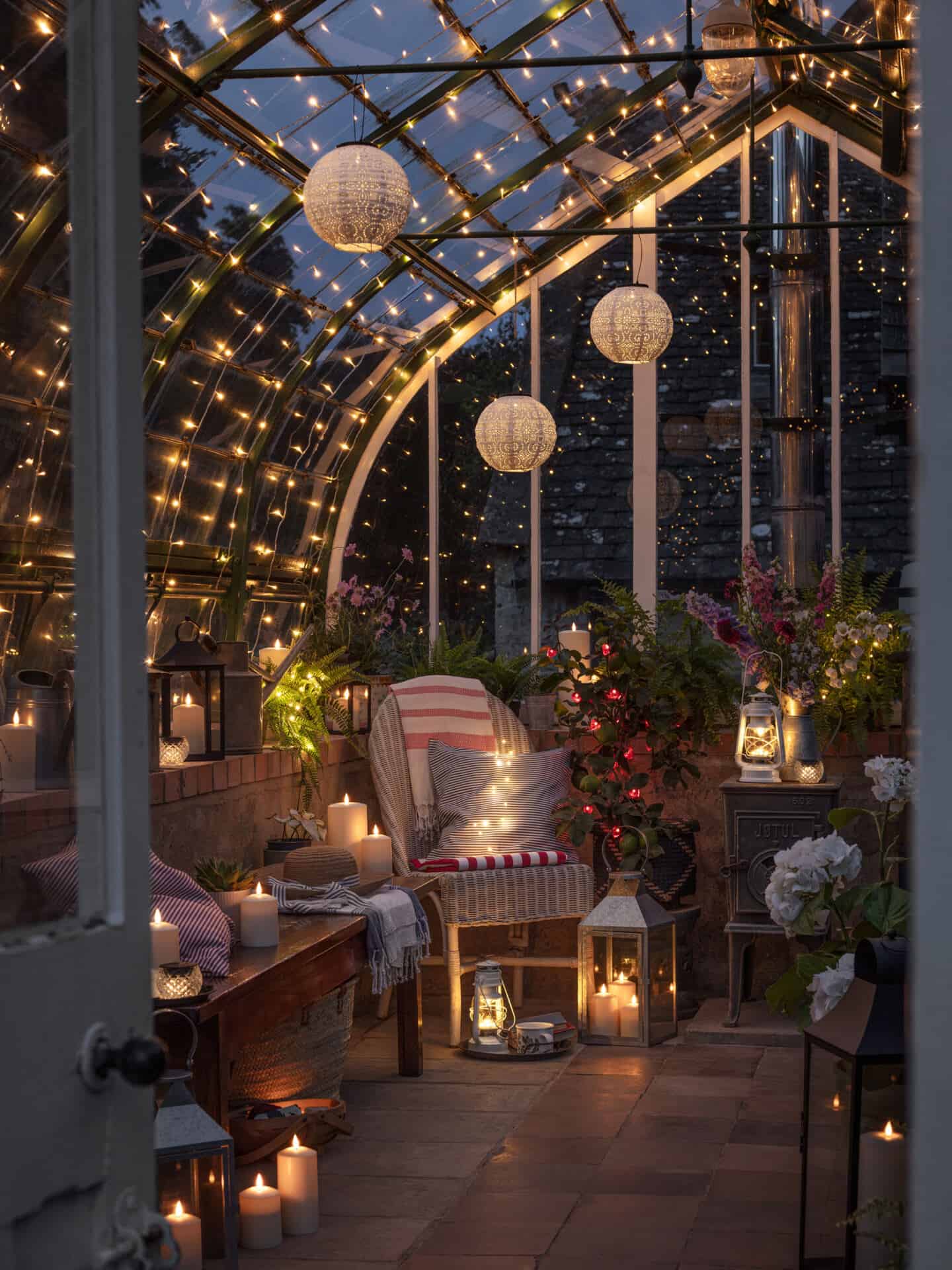 A glass greenhouse is lit up with string lights, lanterns and candles