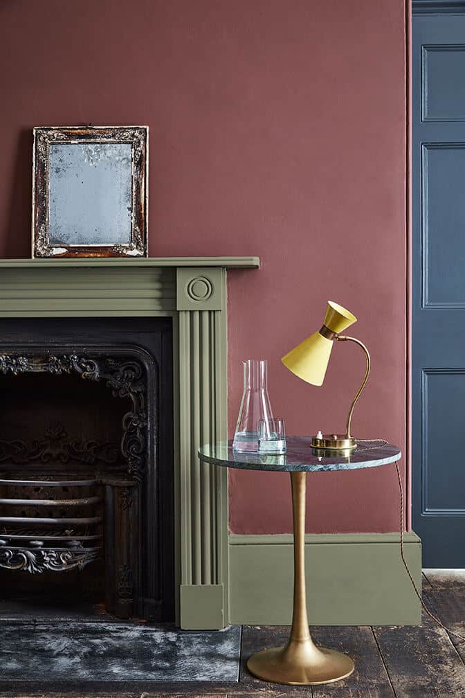 A close up of an old iron fireplace with a green surround and green skirting boards agaist a pink wall with a blue door in the background. Image c/o Little Greene