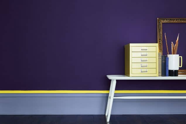 A white desk with filing cabinet on top in front of a purple wall. The skirting board has a neon yellow stripw at the top. Image c/o Little Greene