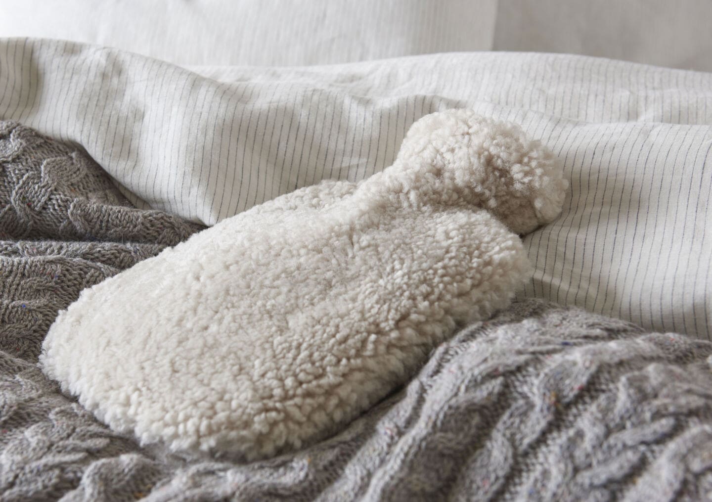 A hot water bottle in a a fluffy white cover laid on a grey woollen blanket on  a bed