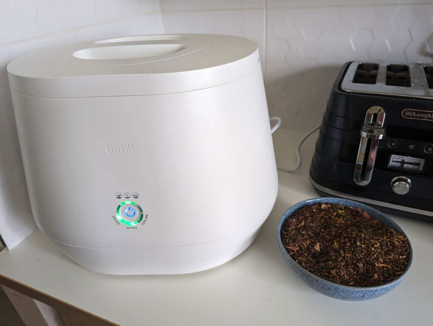 Lomi kitchen composter on a worktop next to a bowl of Lomi compost and a toaster