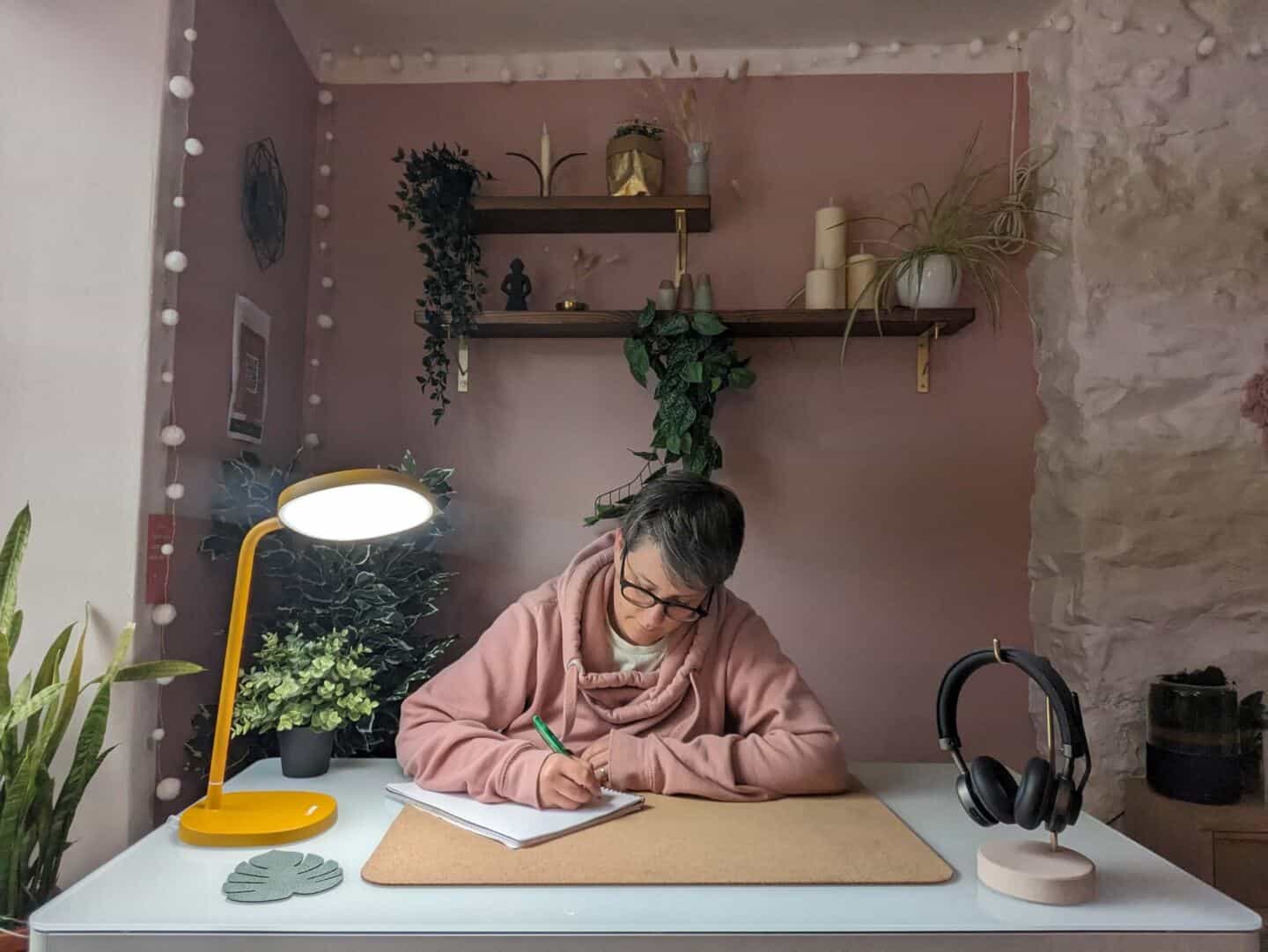 A woman in her forties sat at a desk writing in the light of the Lumie Task lamp