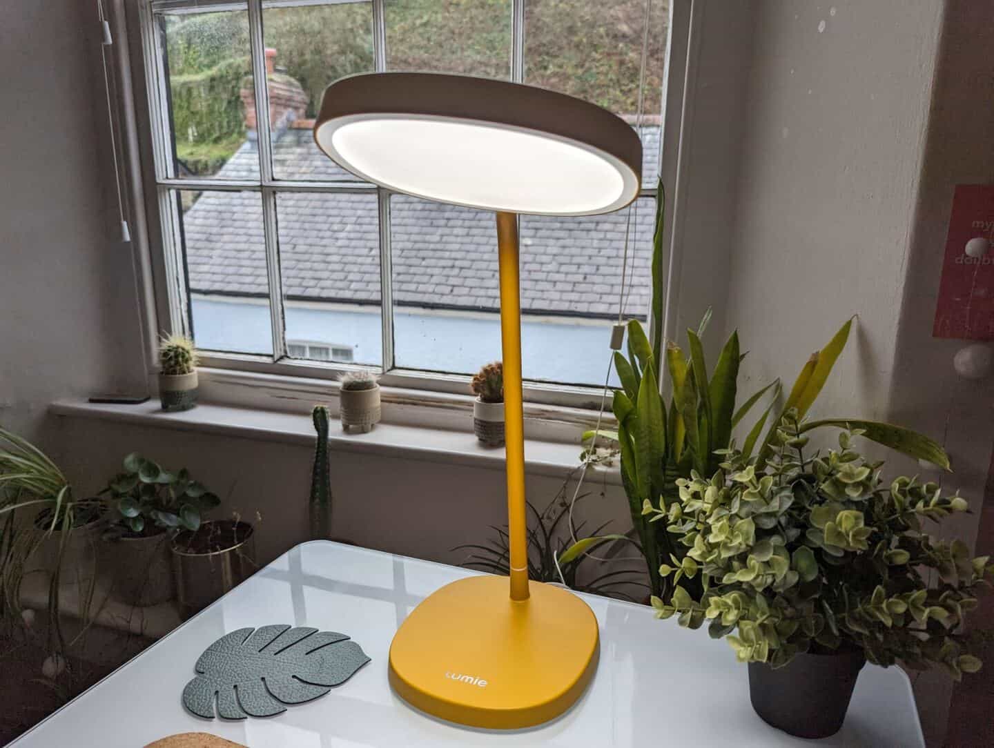 The Lumie Task lamp on a white desk, positioned in front of a window surrounded by plants