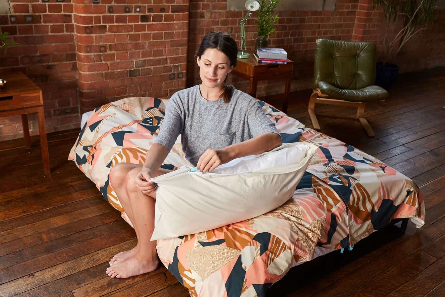 Koa revolutionary duvet cover. A women zips up a pillow case whilst sitting on a bed in an industrial loft style bedroom. The easy way to change a duvet cover