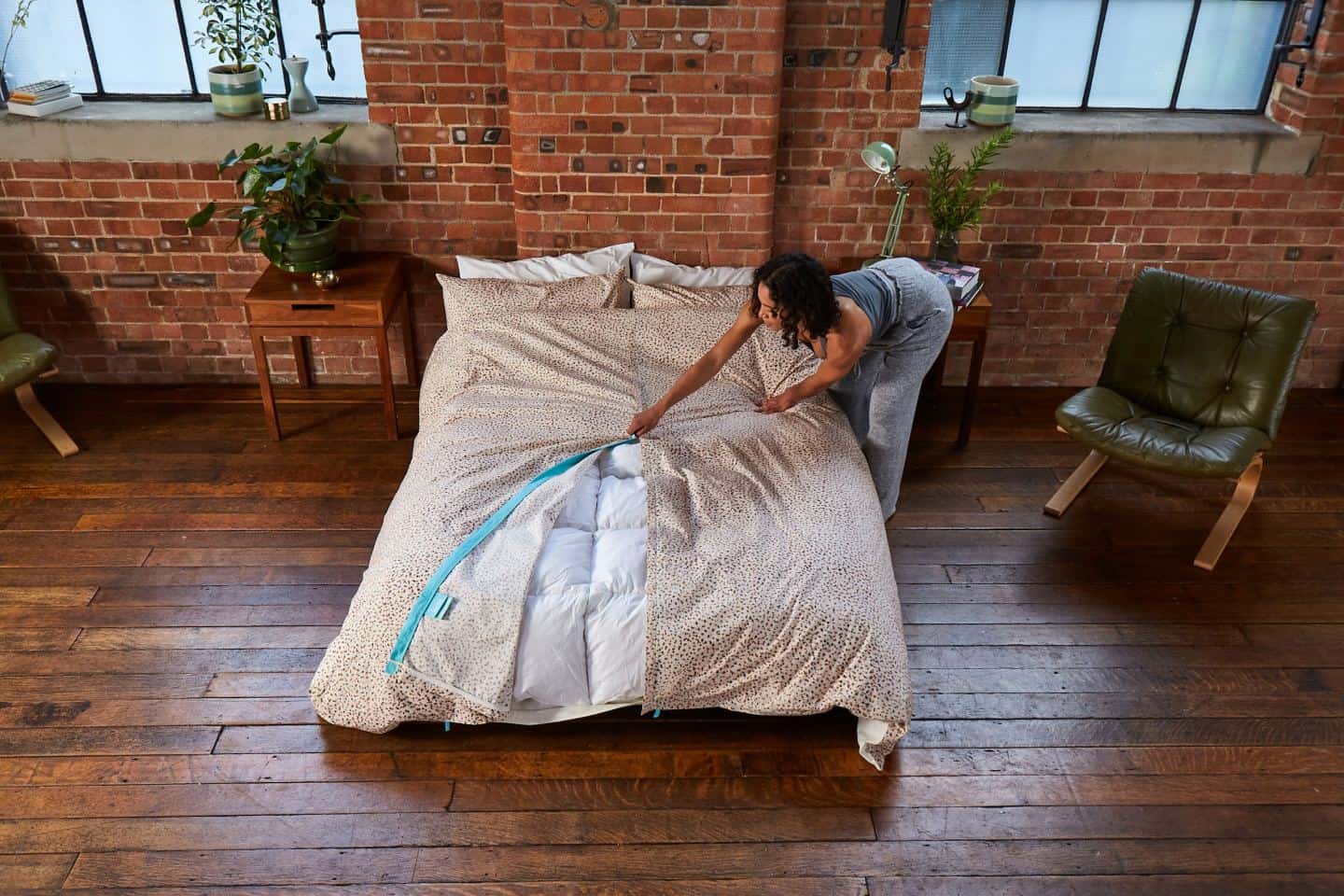 A young black women puts a Koa duvet cover on a bed in an elegant industrial-style bedroom with wood floors and red brick walls